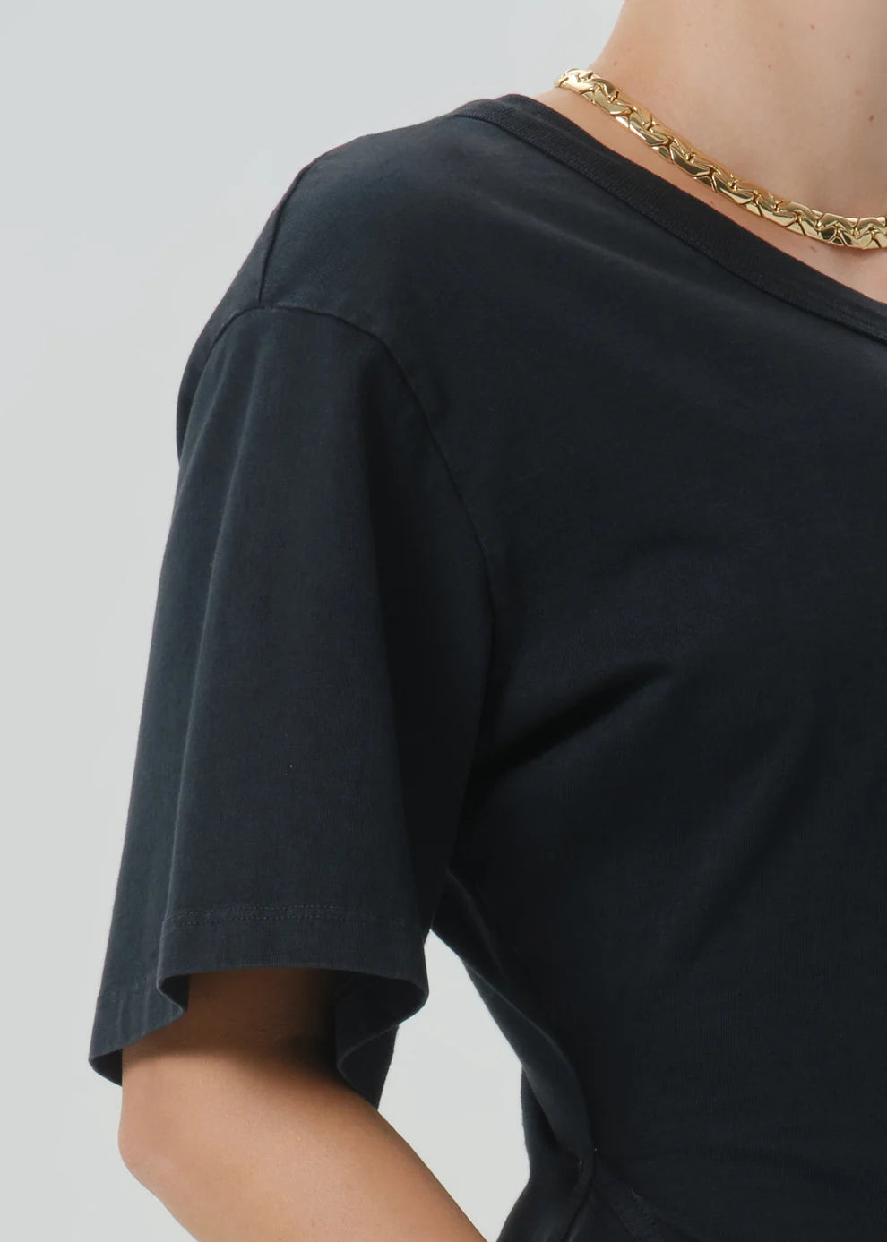Close-up of a person wearing a black short-sleeve crew-neck T-shirt and a chunky gold chain necklace against a light gray background.