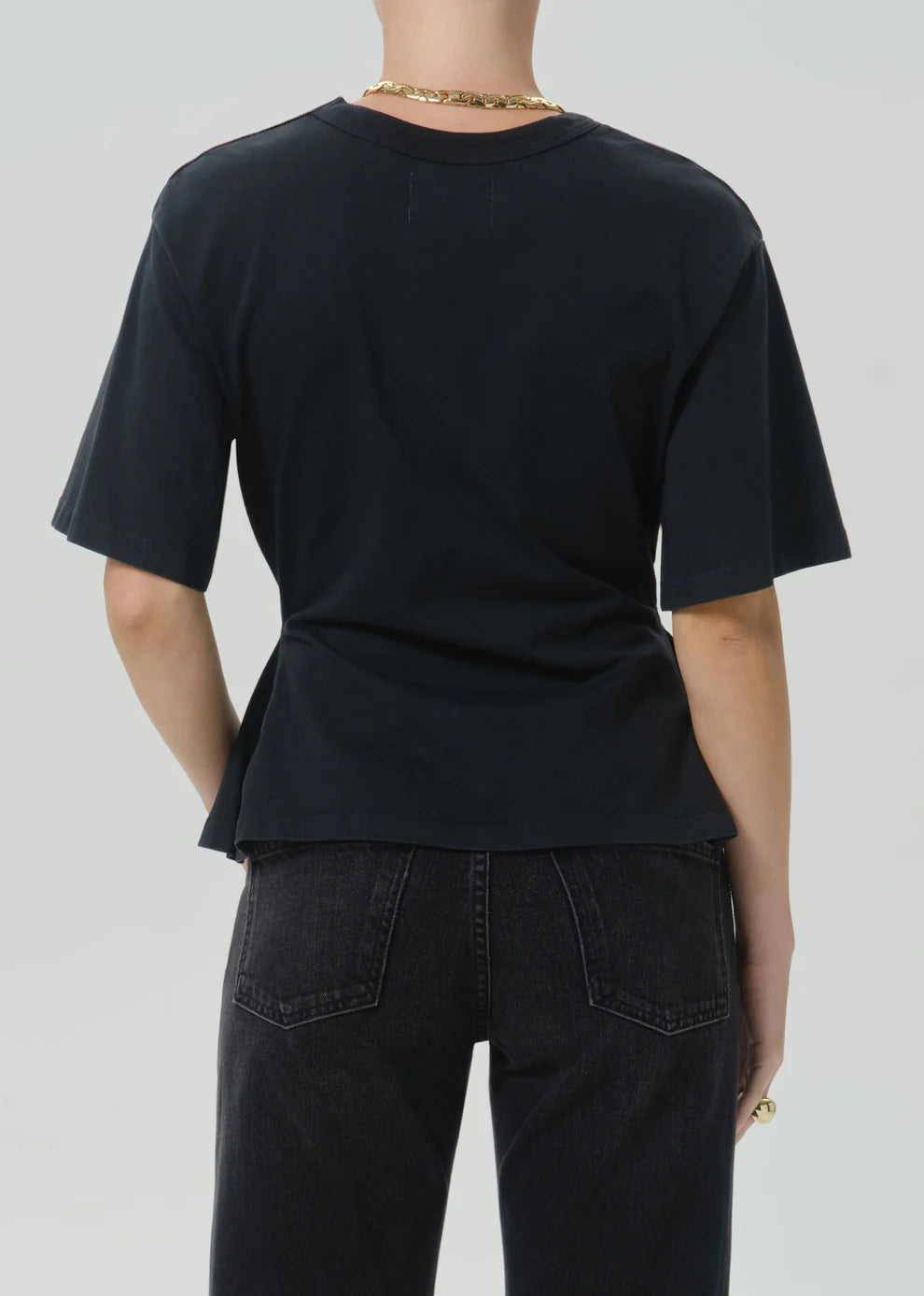Rear view of a person wearing a black short-sleeve crew-neck T-shirt and dark jeans against a light gray studio background.