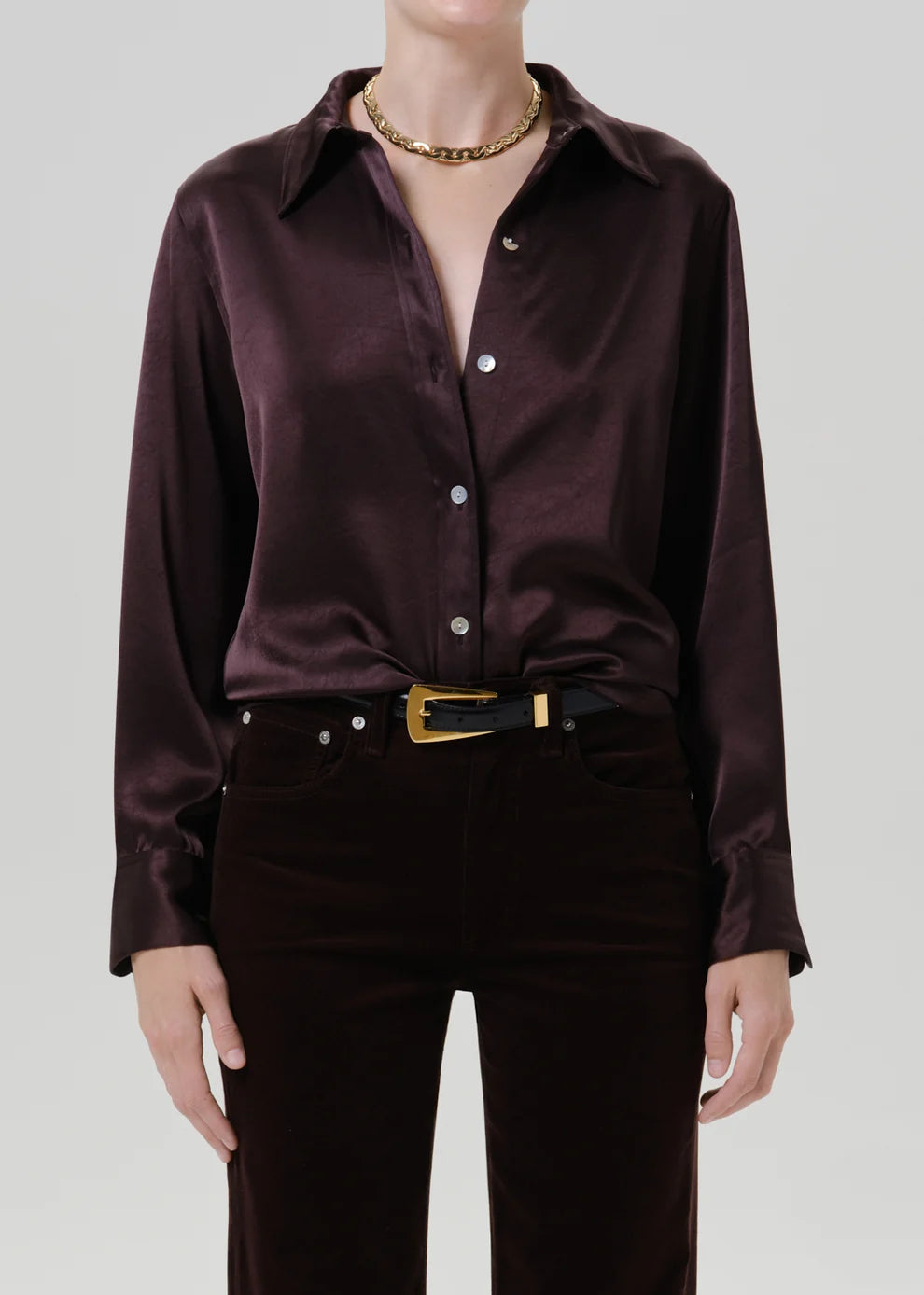 A model wearing a dark brown satin button-down shirt tucked into dark brown corduroy trousers with a gold belt, against a light gray studio background.