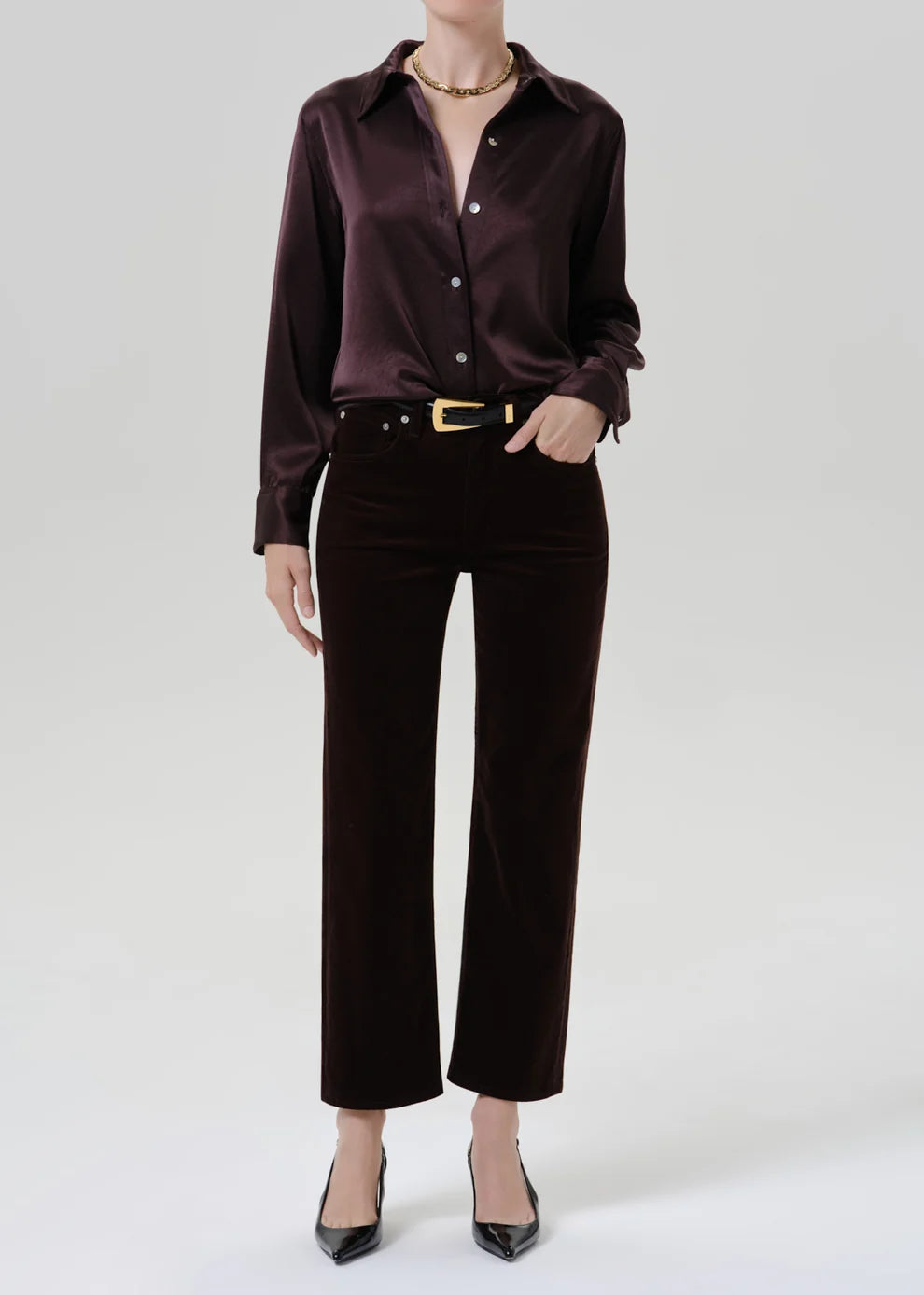 A model wearing a dark brown satin long-sleeve button-up shirt and matching dark brown velvet cropped trousers, styled with a gold belt, necklace, and black pointed-toe heels against a light gray studio background.