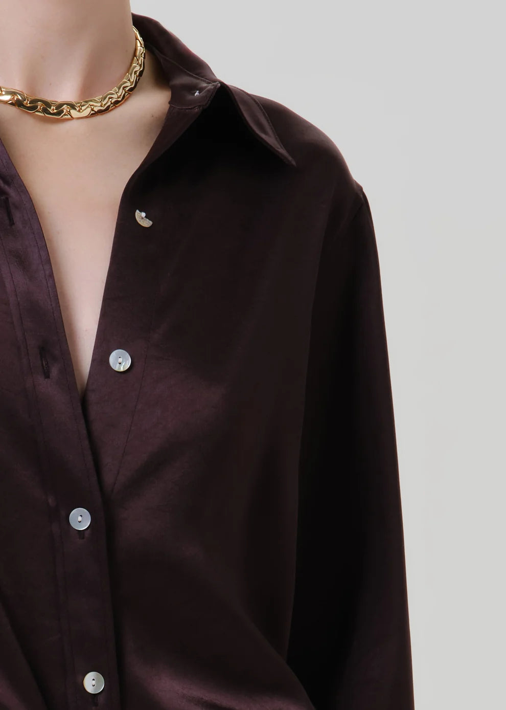 Close-up of a person wearing a dark brown satin button-up shirt with silver buttons and a chunky gold chain necklace, against a light gray background.
