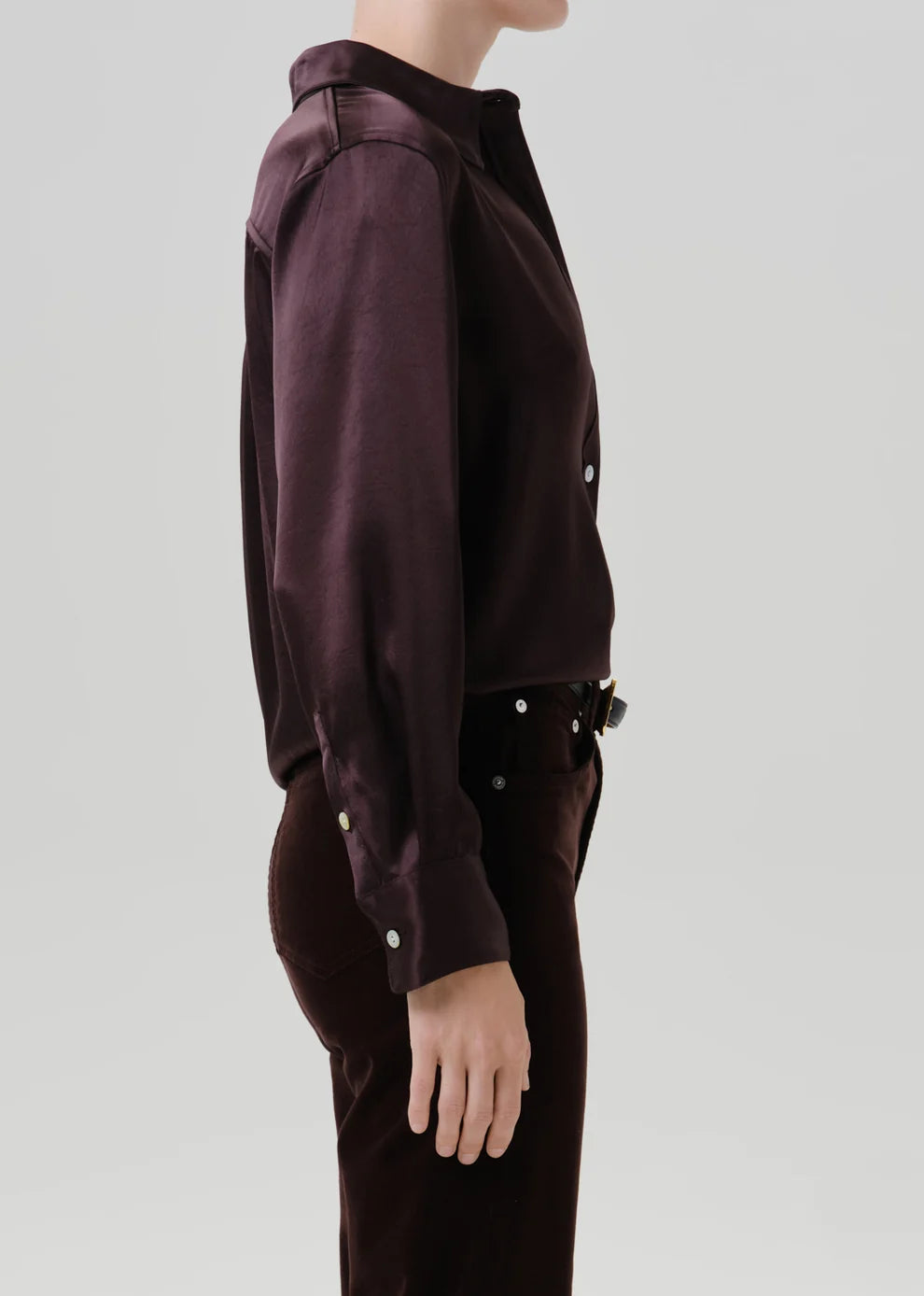 A side-profile view of a model wearing a dark plum button-up shirt and matching dark trousers against a light gray studio background.