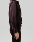 A side-profile view of a model wearing a dark plum button-up shirt and matching dark trousers against a light gray studio background.