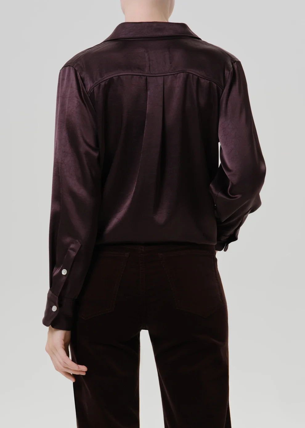 Rear view of a model wearing a dark plum satin button-up shirt and matching dark trousers against a light gray studio background.