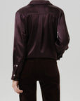 Rear view of a model wearing a dark plum satin button-up shirt and matching dark trousers against a light gray studio background.