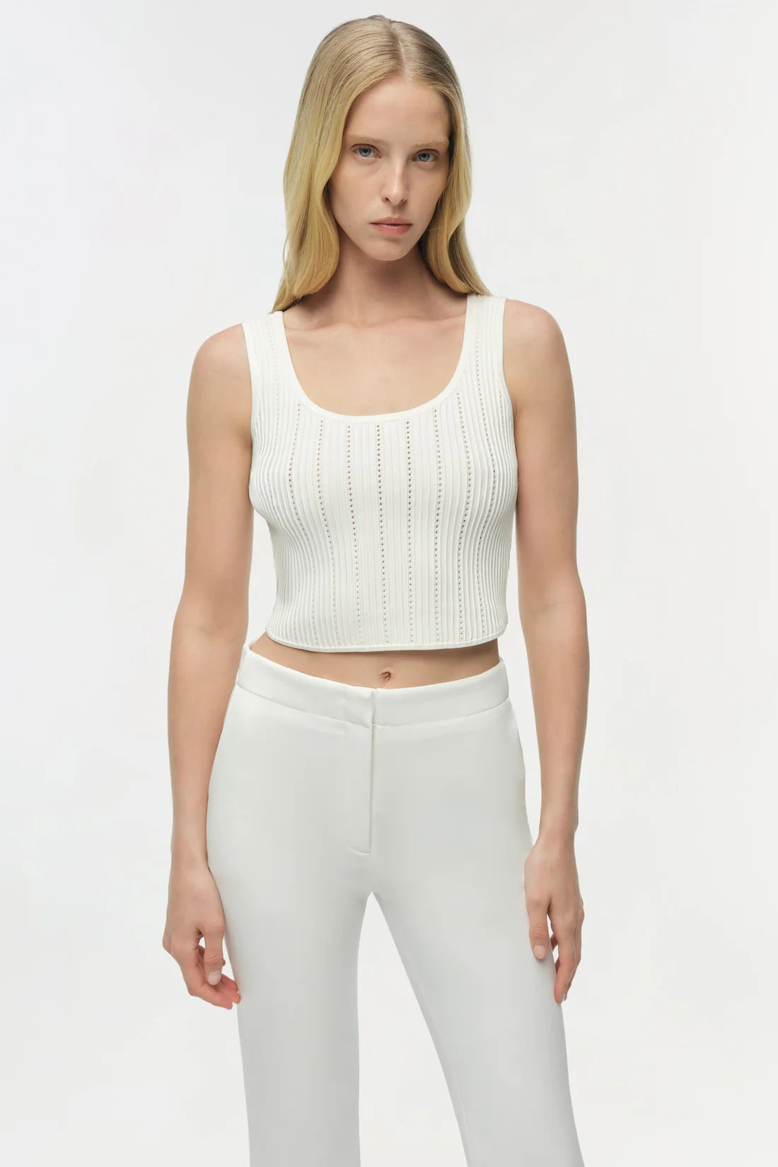 Woman wearing a white ribbed tank top and white pants on a plain background