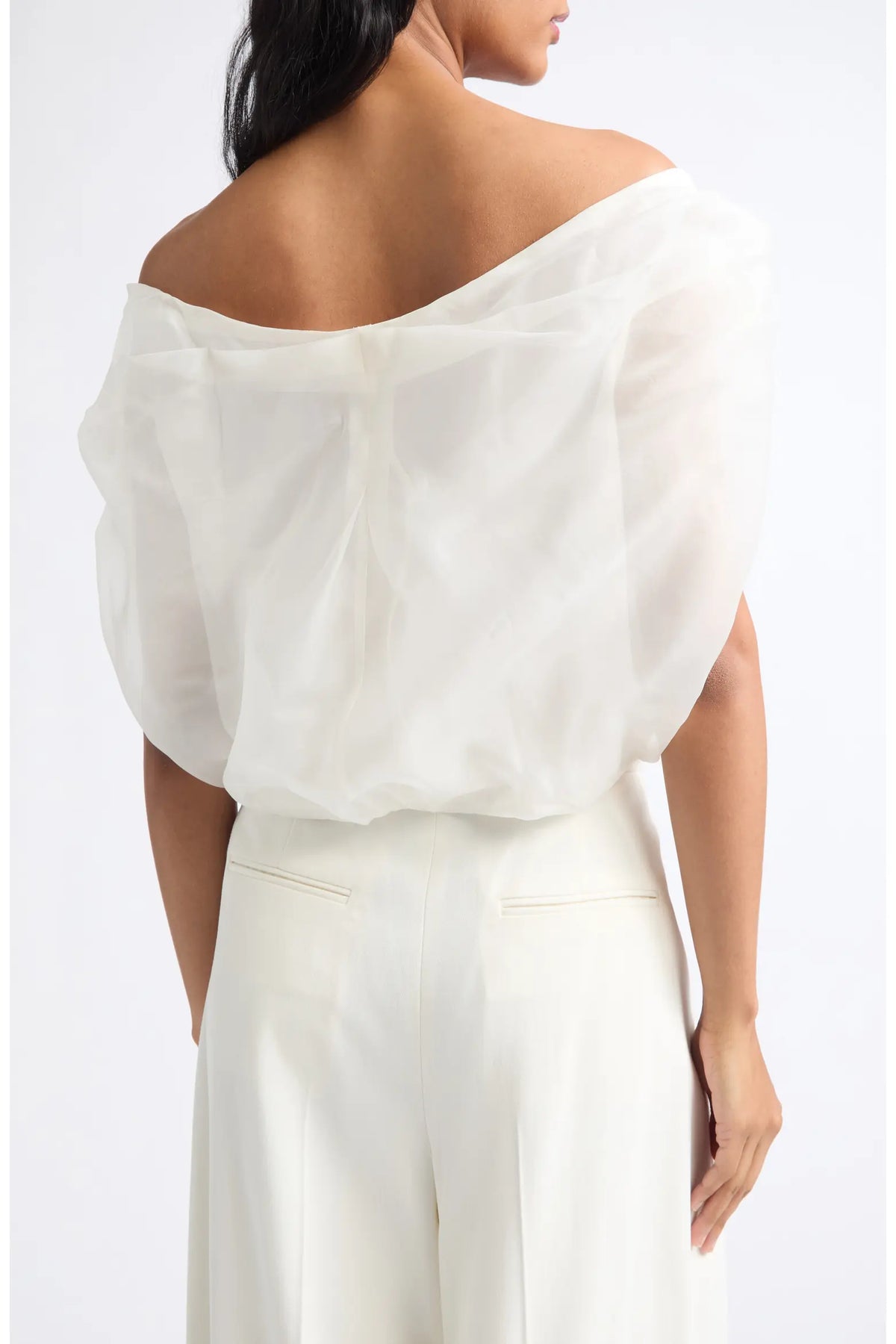 A woman shown from the back wearing a white off-the-shoulder sheer puff-sleeve blouse and white wide-leg trousers against a plain light background.