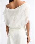 A woman shown from the back wearing a white off-the-shoulder sheer puff-sleeve blouse and white wide-leg trousers against a plain light background.
