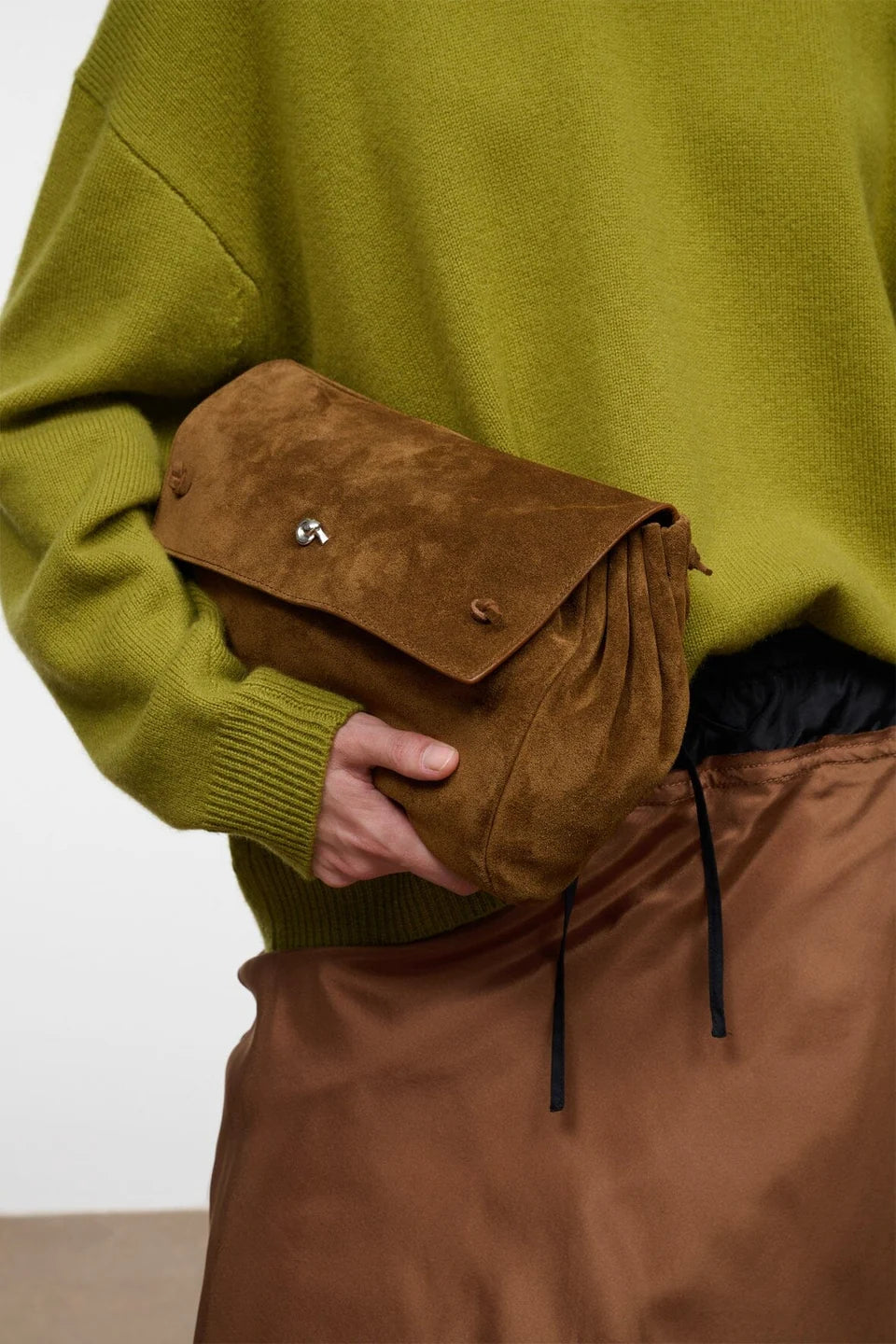 A person wearing an olive green sweater and brown skirt holds a brown suede flap pouch bag with a silver-tone metal clasp.