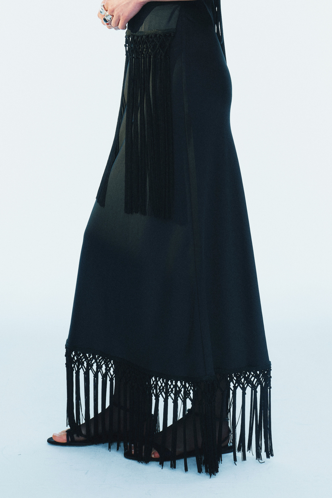 model wearing a black maxi skirt with fringe trim on a white background, side view