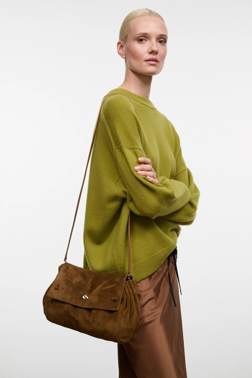 A model wearing an olive green knit sweater and brown pants, carrying a brown suede shoulder bag with a small silver clasp against a plain light gray background.
