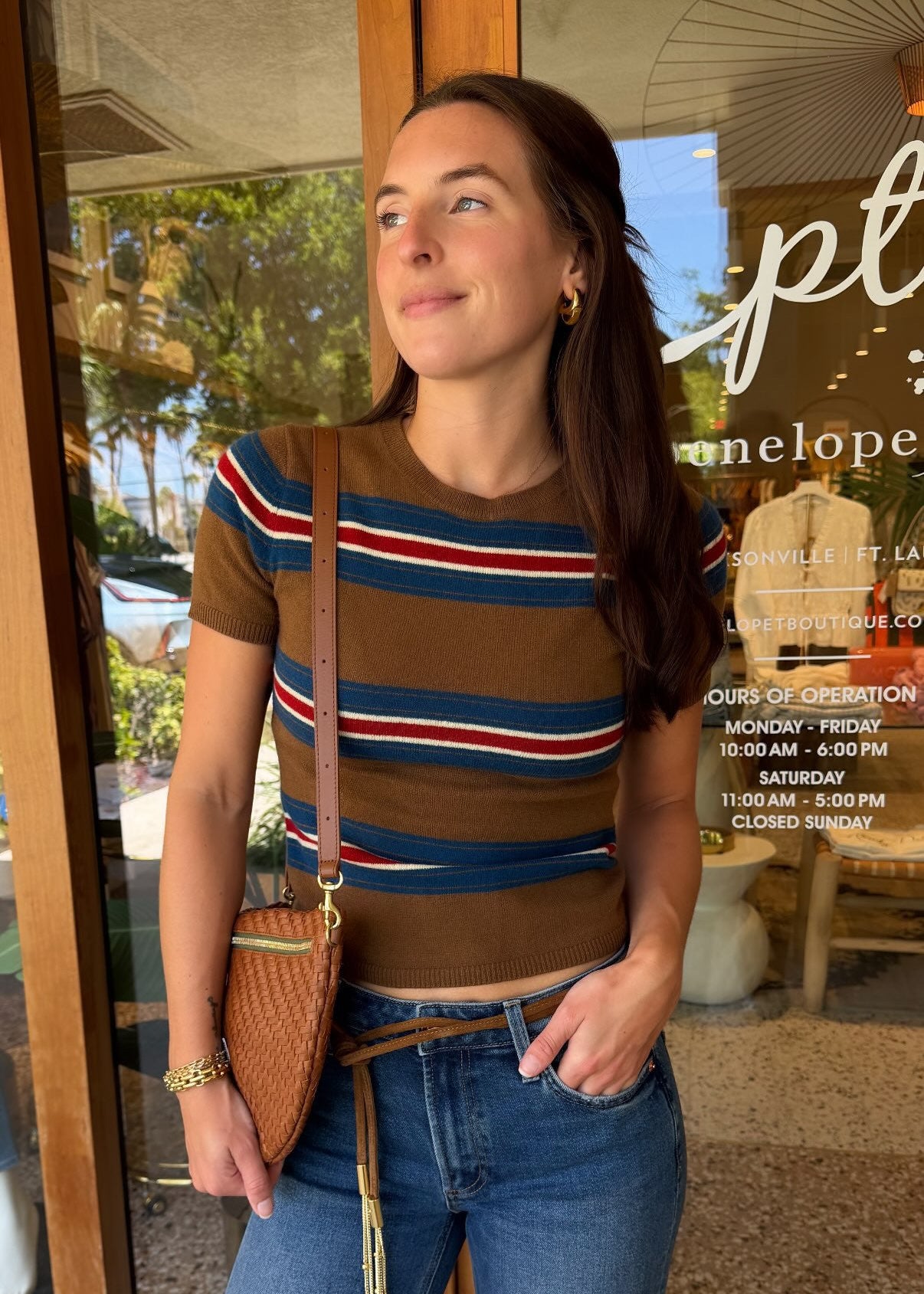 A woman wearing a brown short-sleeve crop top with red and blue horizontal stripes, paired with blue jeans and a brown woven crossbody bag, standing in front of a storefront window.