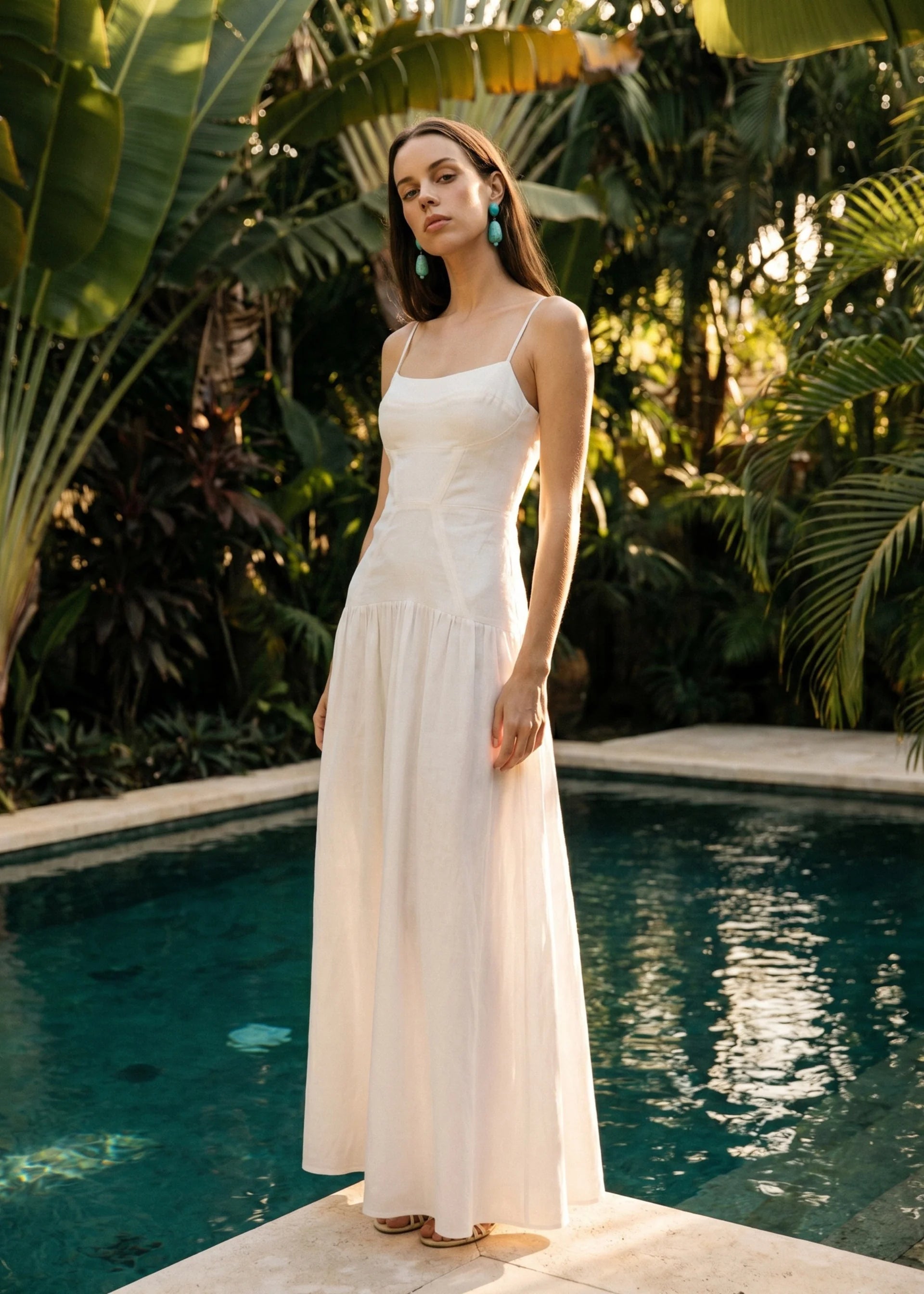 Woman in a white dress standing by a pool with lush greenery in the background