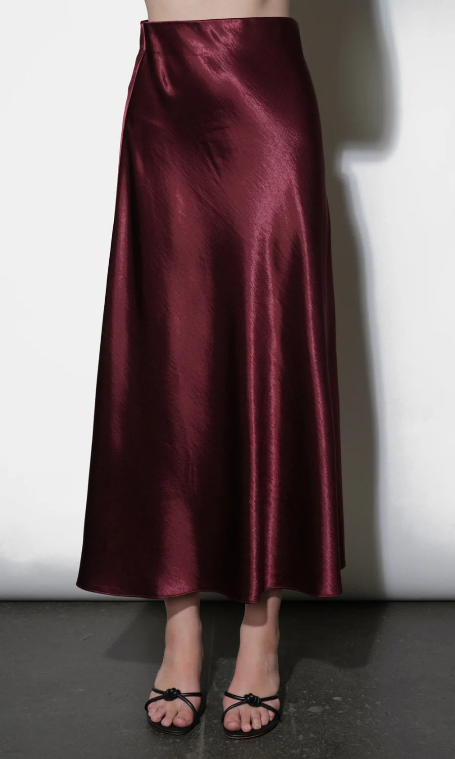A floor-length satin skirt in a deep burgundy red, shown on a model from the waist down against a white wall.
