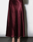A floor-length satin skirt in a deep burgundy red, shown on a model from the waist down against a white wall.