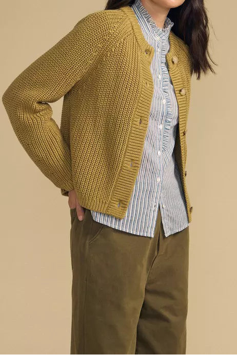 A woman modeling a mustard yellow ribbed knit cardigan over a blue and white striped button-down shirt and olive green pants, set against a solid beige studio background.