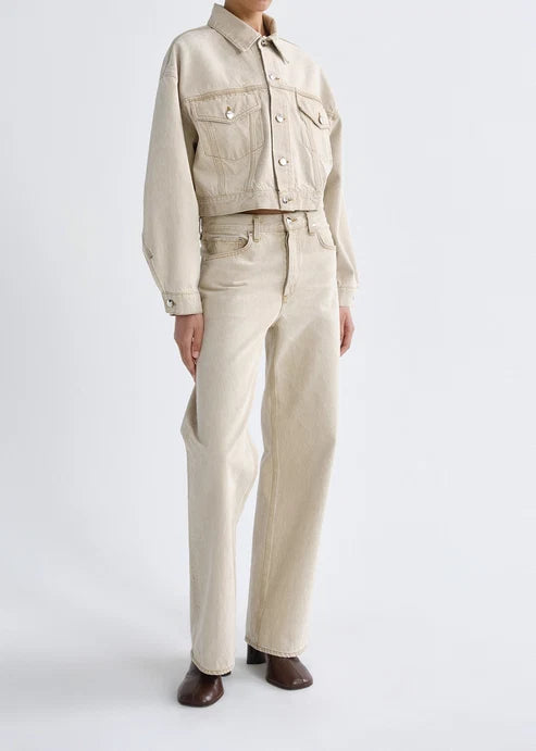 A model wearing a light beige cropped denim jacket and matching straight-leg jeans, paired with dark brown leather shoes, against a plain light gray studio background.