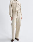 A model wearing a light beige cropped denim jacket and matching straight-leg jeans, paired with dark brown leather shoes, against a plain light gray studio background.