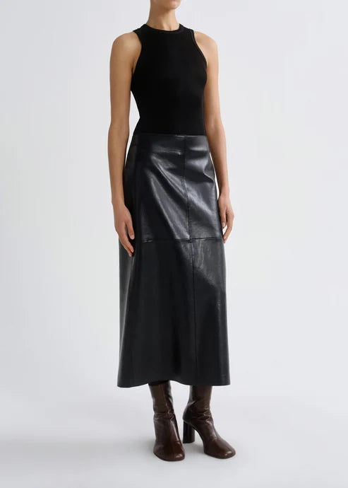 A model wearing a sleeveless black mock-neck top and a black faux-leather midi skirt, paired with dark brown heeled boots, against a plain light gray studio background.