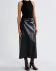 A model wearing a sleeveless black mock-neck top and a black faux-leather midi skirt, paired with dark brown heeled boots, against a plain light gray studio background.