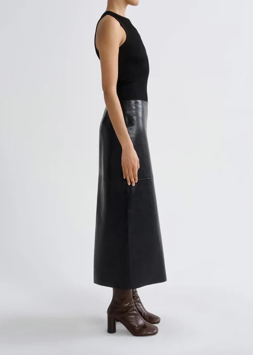 A model shown from the side wearing a black sleeveless knit top and a long black faux-leather midi skirt with a high waist, paired with dark brown heeled ankle boots, against a light gray studio background.