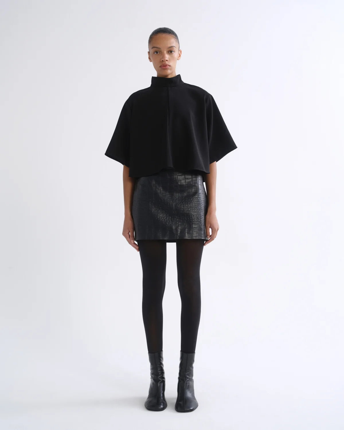 A model stands against a plain white studio background wearing a black short-sleeve mock-neck cape top, a black textured mini skirt, black tights, and black ankle boots.