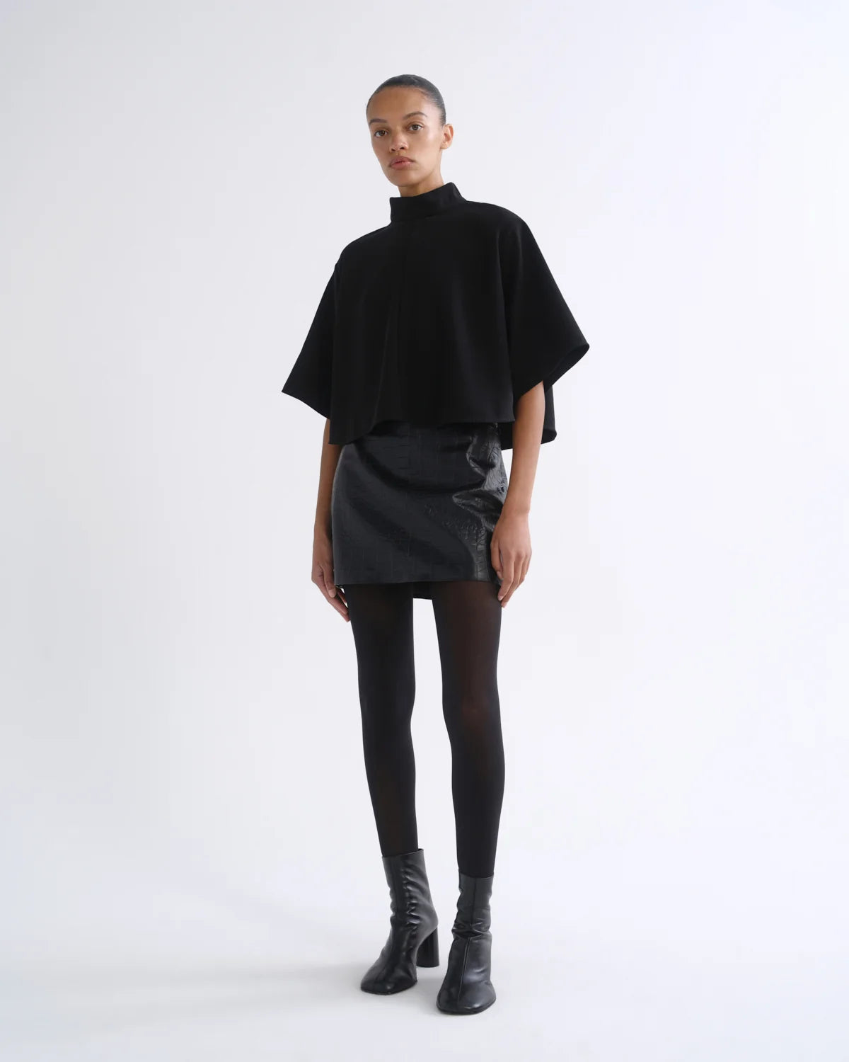 A model stands against a plain white studio background wearing a black high-neck cape-sleeve top, a black mini skirt with tonal side panels, black tights, and black ankle boots.