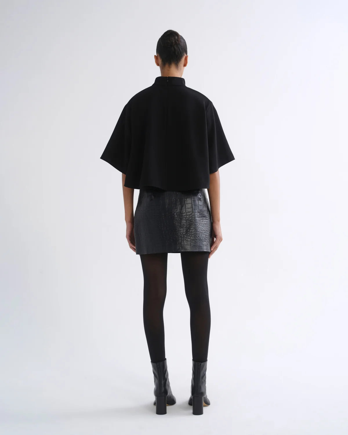 A model viewed from the back wearing a black short-sleeved high-neck top, a textured black mini skirt, black tights, and black ankle boots, set against a plain light gray studio background.