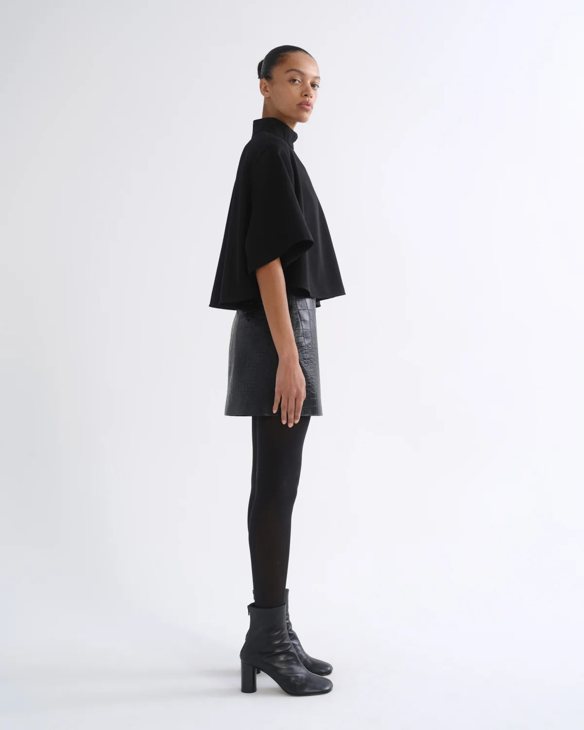 A model is shown in profile wearing a black cropped cape-sleeve top, a black faux-leather mini skirt, black tights, and black heeled ankle boots against a plain white studio background.