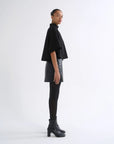 A model is shown in profile wearing a black cropped cape-sleeve top, a black faux-leather mini skirt, black tights, and black heeled ankle boots against a plain white studio background.
