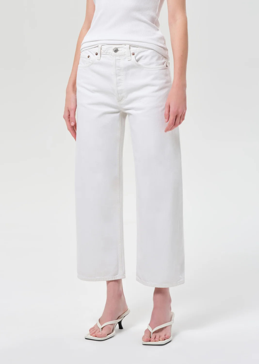 White cropped wide-leg jeans worn by a model, paired with a white top and white heeled thong sandals, photographed against a light gray studio background.