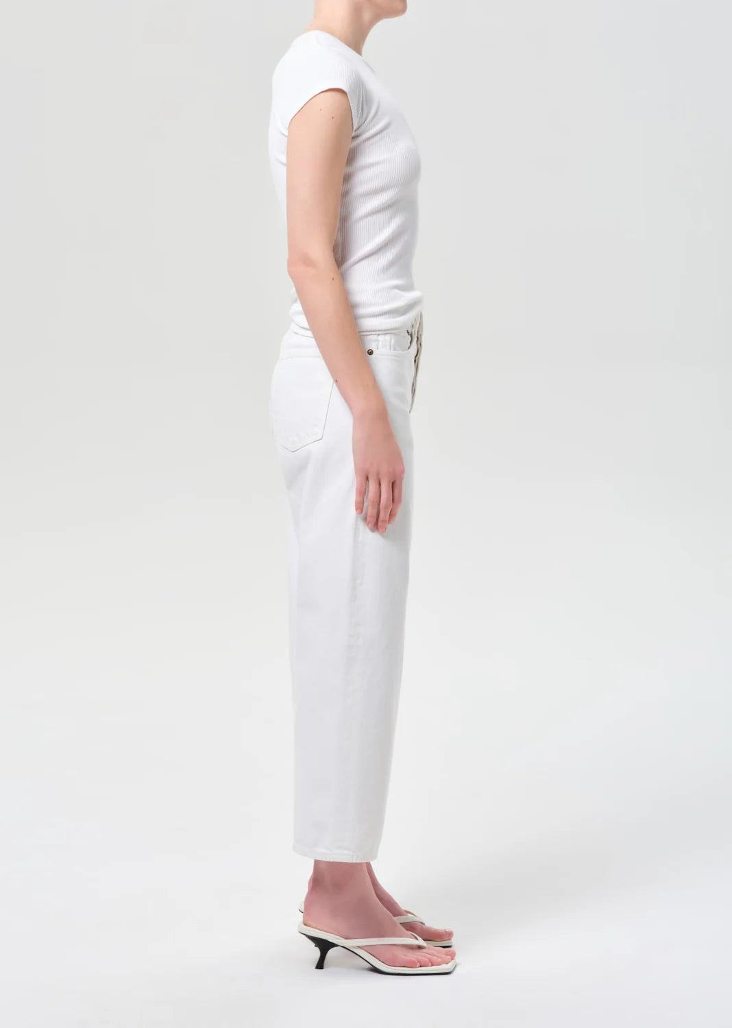 A model shown in profile wearing a white short-sleeve top, white cropped pants, and light pink heeled sandals against a plain light gray studio background.