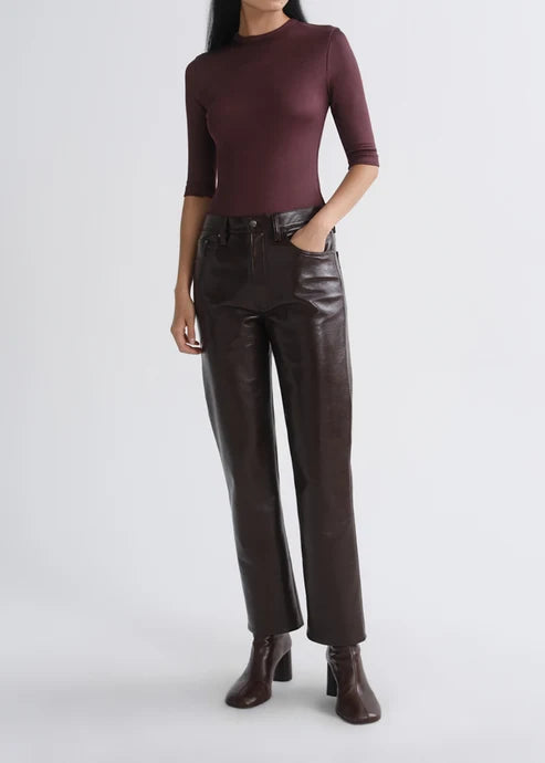 A model wearing a dark burgundy mock-neck long-sleeve top, dark brown leather-look straight-leg pants, and dark brown heeled ankle boots, standing against a light gray studio background.