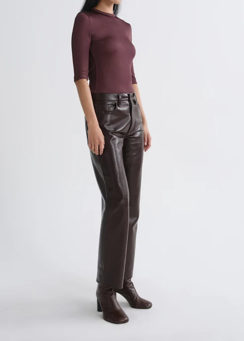 A model wearing a fitted burgundy three-quarter sleeve top, dark brown faux-leather straight-leg pants, and dark brown heeled ankle boots, photographed against a light gray studio background.
