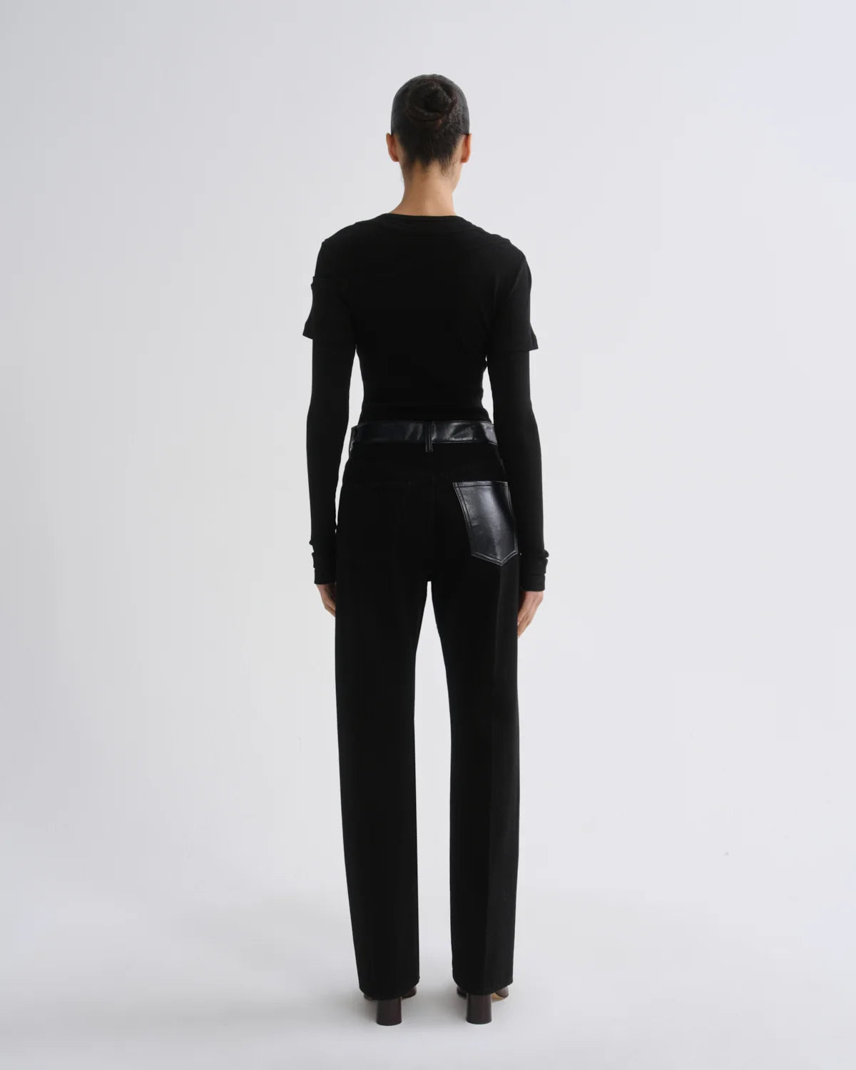 A model viewed from the back wearing a black long-sleeve top with shoulder details and black wide-leg trousers featuring a glossy black leather patch pocket on the right side, set against a plain light gray studio background.