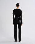 A model viewed from the back wearing a black long-sleeve top with shoulder details and black wide-leg trousers featuring a glossy black leather patch pocket on the right side, set against a plain light gray studio background.