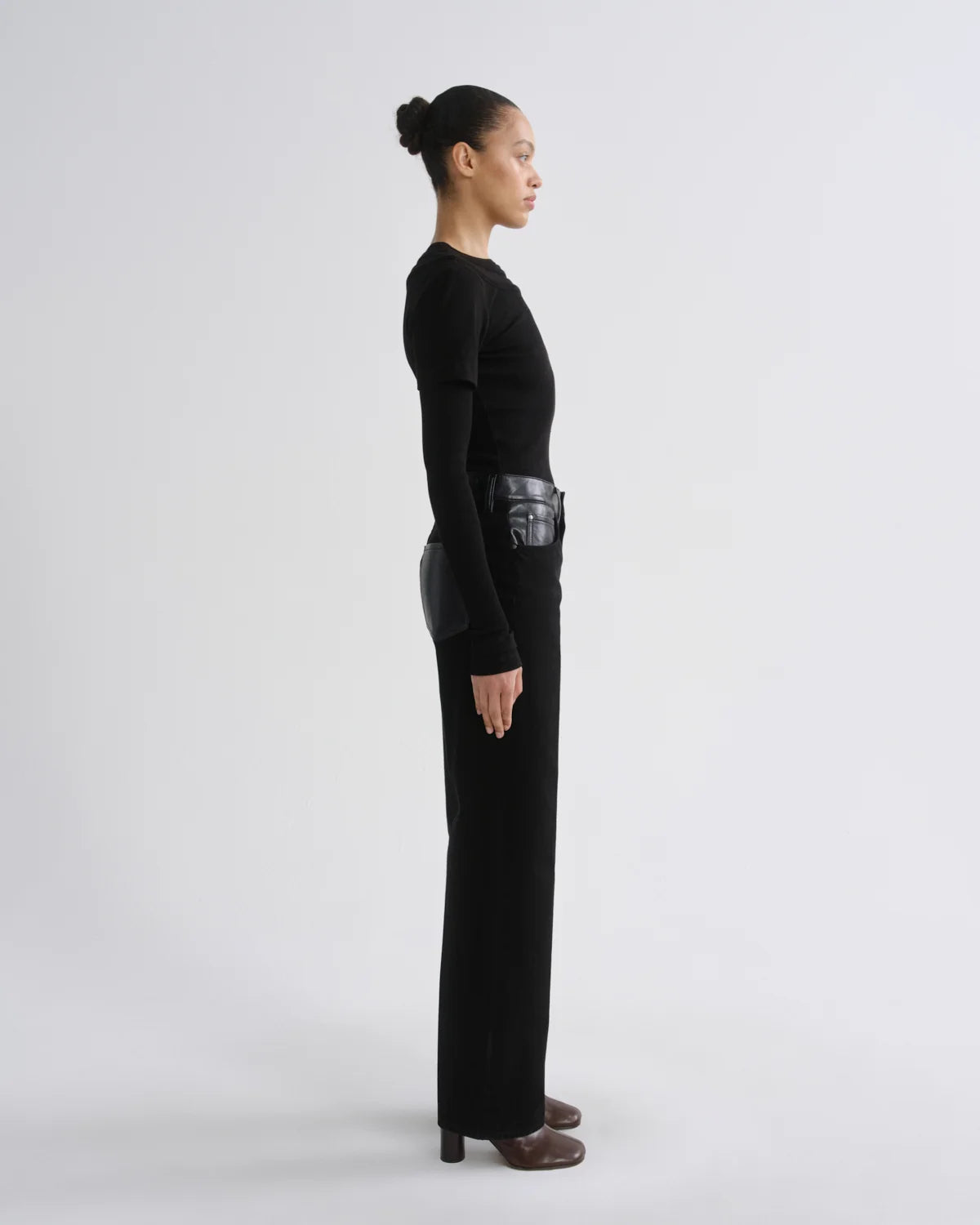 A model in profile wearing a black long-sleeve knit top and matching wide-leg trousers, styled with brown heeled shoes, against a plain light gray studio background.