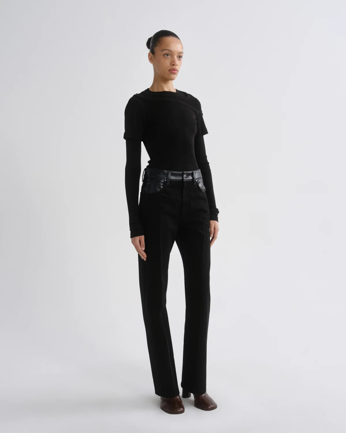 A model stands in a studio wearing a black long-sleeve top, black high-waisted straight-leg pants and brown pointed shoes against a light gray background.