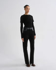 A model stands in a studio wearing a black long-sleeve top, black high-waisted straight-leg pants and brown pointed shoes against a light gray background.