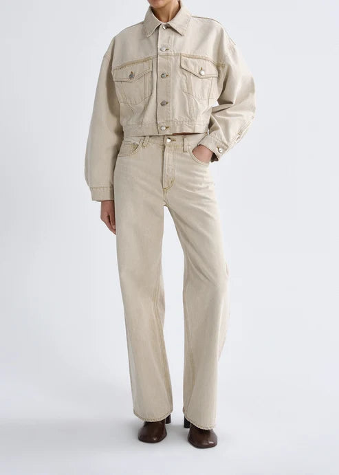 A model wearing a light beige cropped denim jacket and matching wide-leg jeans, paired with dark brown shoes, against a plain light gray studio background.
