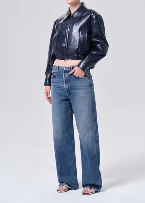 A model wearing a shiny dark navy cropped leather jacket and wide-leg blue jeans, standing against a light gray studio background.