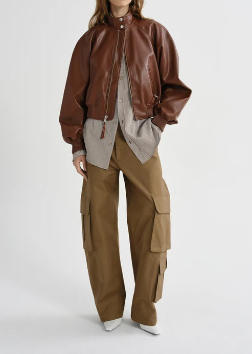 A model wearing a brown leather bomber jacket over a light gray shirt, paired with wide-leg brown cargo pants and white pointed shoes, against a light gray studio background.