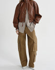 A model wearing a brown leather bomber jacket over a light gray shirt, paired with wide-leg brown cargo pants and white pointed shoes, against a light gray studio background.