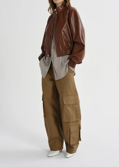 A model wearing a brown leather bomber jacket, a light gray shirt, brown cargo pants, and white pointed shoes, photographed against a plain light gray studio background.