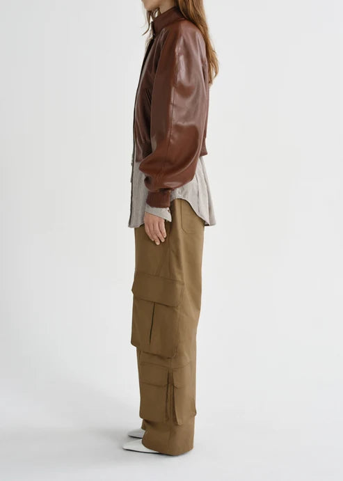 A model shown from the side wearing a brown faux-leather jacket, a light beige shirt, and wide-leg brown cargo pants against a plain light gray background.