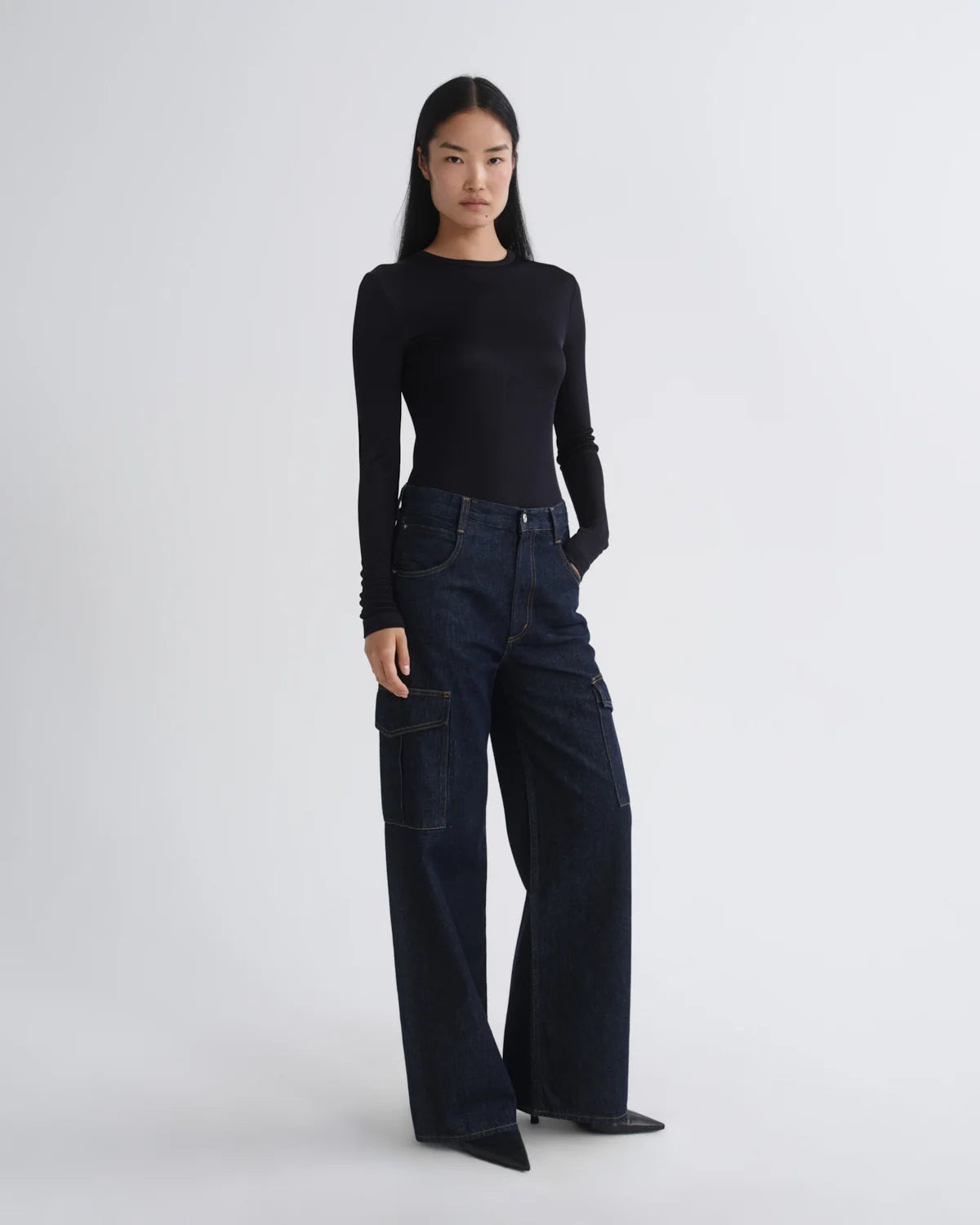 A model stands against a light gray studio background wearing a black long-sleeve top and dark wide-leg denim cargo pants.