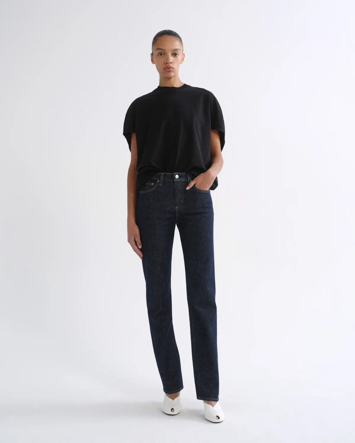 A model stands against a plain white studio background wearing a black short-sleeve t-shirt, dark-wash straight-leg jeans, and white pointed shoes.
