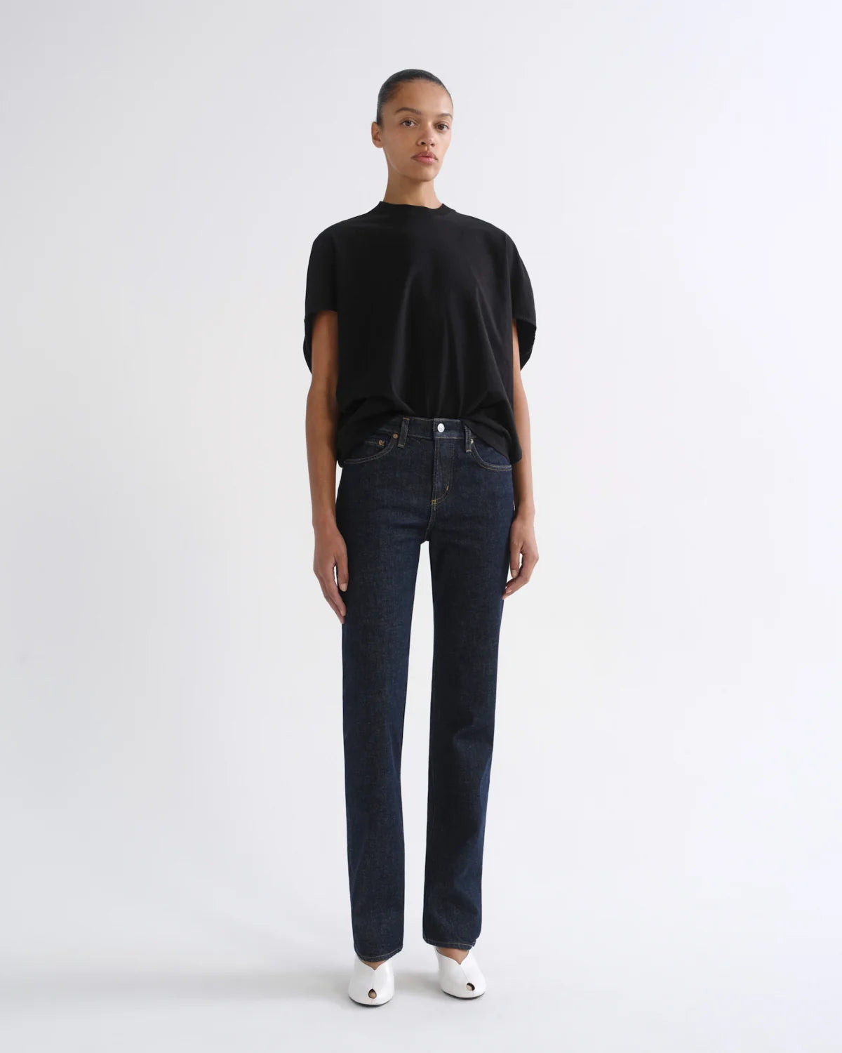 A model stands against a plain white studio background wearing a black short-sleeve t-shirt, dark blue straight-leg jeans, and white pointed shoes.