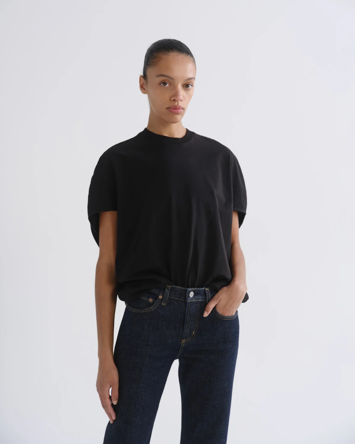 A model wearing a black short-sleeve crewneck t-shirt tucked into dark blue jeans, standing against a light gray studio background.