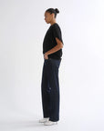 A model shown in profile wearing a black short-sleeve t-shirt, dark blue wide-leg denim jeans, and white pointed-toe heeled shoes, standing against a plain white studio background.